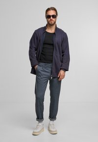 Navy Zip-Up-Jacke über einem schwarzen Tanktop, kombiniert mit hellblauen gemusterten Hosen und weißen Sneakers, vor einem grauen Hintergrund stehend.