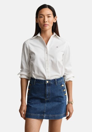 Donna che indossa una camicia bianca con bottoni e le maniche arrotolate e una gonna di jeans blu con bottoni dorati ai lati, in piedi davanti a uno sfondo semplice.