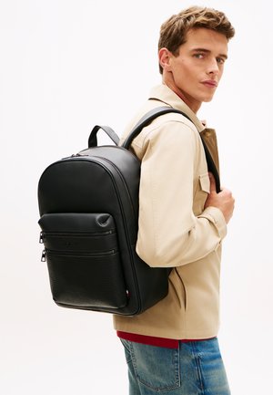 Jauns vīrietis, tērpies bēšā jaku un zilas džinsās, pār vienu plecu nēsā lielu melnu mugursomu un skatās pār labo plecu.