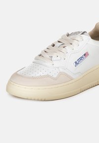 Baskets blanches en cuir avec des accents en suède beige, boîtier perforé à l'avant, semelle en caoutchouc crème et marque visible sur le côté.