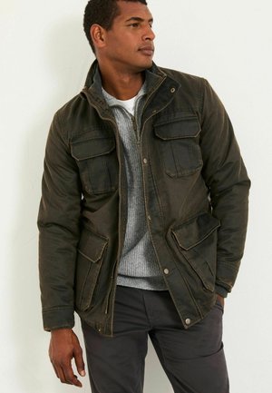 Olive green, waxed fabric jacket with a zippered front, four front pockets, and buttoned cuffs. Worn over a gray sweater, with a relaxed fit.