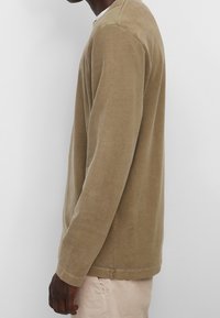 Langarmshirt aus beigem Baumwollstoff, mit abgerundetem Saum, lässiger Passform und detaillierten Nähten an der Schulter. Weiche Textur und minimales Design.