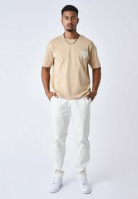 Camiseta de algodón beige con mangas cortas y parche de corazón en el lado izquierdo del pecho, combinada con pantalones blancos y zapatillas blancas.
