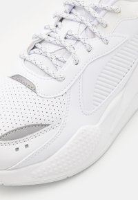 Chaussure de sport blanche avec embout perforé, semelle texturée, panneaux cousus et lacets blancs à motifs sur un fond clair.