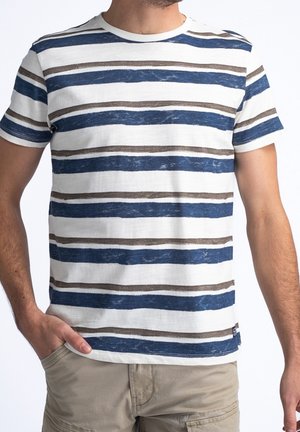 Mann mit weißem T-Shirt mit unregelmäßigen blauen und braunen horizontalen Streifen und beigefarbener Hose, eine Hand in der Tasche.