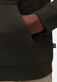 Sudadera de color verde oscuro con un bolsillo tipo canguro, puños y dobladillo acanalados. Textura suave con detalles de costura y una pequeña etiqueta con el logo en el lateral.