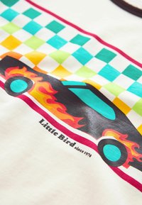 Hvid t-shirt med et farverigt bilgrafik med flammer, ternet mønster i teal, grøn, orange og rød, samt teksten "Little Bird siden 1974".