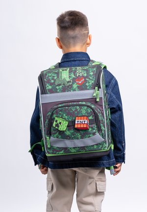 Kind trägt einen grünen Tarn-Minecraft-Rucksack mit Creeper- und TNT-Designs, bekleidet mit einer blauen Jacke und beigefarbenen Hosen, von hinten gesehen.