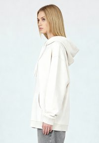 Sudadera con capucha oversize en color crema, con bolsillo frontal, capucha con cordón ajustable y puños acanalados. Tejido suave y ajuste relajado.