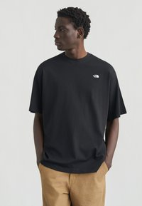 Νεαρός με κοντά σγουρά μαλλιά, φορώντας μαύρο T-shirt The North Face και καφέ παντελόνι, με τα χέρια στις τσέπες, κοιτάζει προς τα αριστερά σε λευκό φόντο.