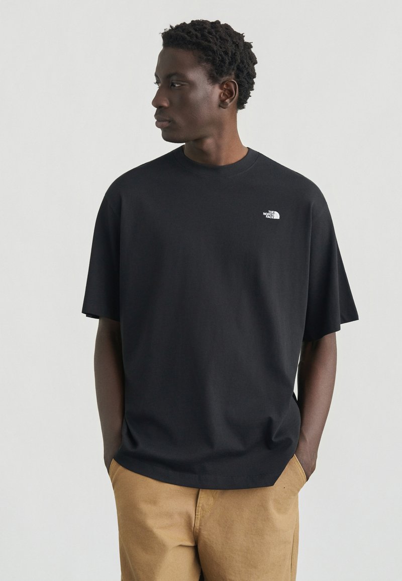 Νεαρός με κοντά σγουρά μαλλιά, φορώντας μαύρο T-shirt The North Face και καφέ παντελόνι, με τα χέρια στις τσέπες, κοιτάζει προς τα αριστερά σε λευκό φόντο.