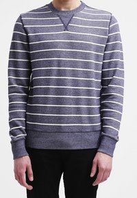 Man in een marineblauwe trui met ronde hals en witte horizontale strepen, met zwarte broek, staand tegen een witte achtergrond.