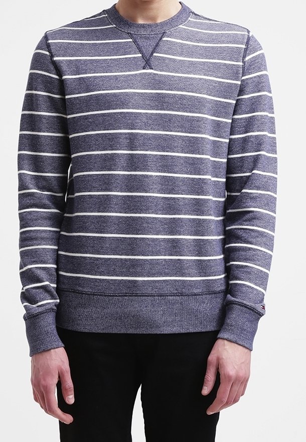 Man in een marineblauwe trui met ronde hals en witte horizontale strepen, met zwarte broek, staand tegen een witte achtergrond.