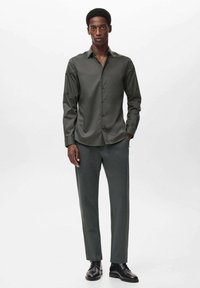 Camicia a maniche lunghe verde scuro con chiusura a bottoni, abbinata a pantaloni coordinati e scarpe nere, esibita su un modello su uno sfondo chiaro.