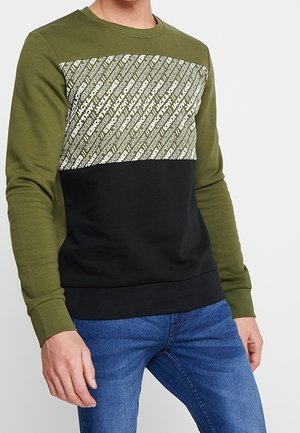 Sudadera verde oliva y negra con un panel frontal blanco con patrón. Tiene un cuello redondo y puños de canalé. Textura de tela suave.