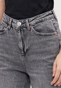 Aukštai užsisegtos juodos ir baltos džinsinės kelnės su blukusia tekstūra, penkiomis kišenėmis, variniais prisuktais taškais ir sidabriniu mygtuko užsegimu.