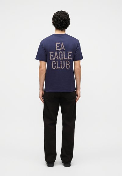 Άτομο με σγουρά μαλλιά, γυρισμένο πλάτη, φορώντας μπλε ναυτικό μπλουζάκι με το κείμενο «EA EAGLE CLUB» και μαύρο παντελόνι, σε απλό λευκό φόντο.