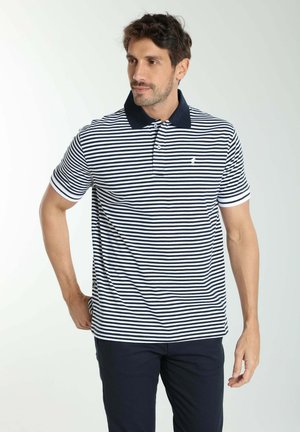 Homme portant un polo à rayures horizontales bleu marine et blanc avec un col sombre et un pantalon sombre, debout avec une main sur la hanche.