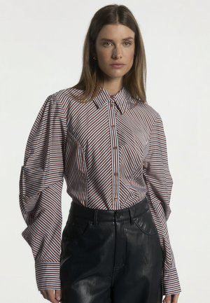 Vrouw draagt een bruin, wit en zwart gestreept overhemd met pofmouwen en een zwarte leren broek, staand tegen een effen achtergrond.