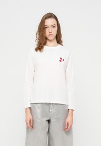 Λευκό μακρυμάνικο πουκάμισο από βαμβάκι, με τρία κόκκινα σχέδια φράουλας στο αριστερό στήθος. Συνδυάζεται με λαμπερό ασημί παντελόνι φαρδιάς γραμμής.