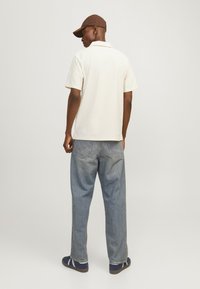 Camisa polo de manga corta en color crema, de material de algodón, vista trasera. Llevada con unos vaqueros azules holgados y zapatillas marinas con rayas blancas y una gorra marrón.