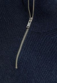 Marineblauer Strickpullover mit einem strukturierten Muster, ausgestattet mit einem silbernen Reißverschluss an der Vorderseite zum Schließen. Weiches Material ist sichtbar.