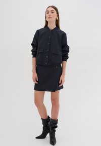 Giacca blu navy con chiusura a bottoni e due tasche frontali, abbinata a una gonna coordinata. Indossata con stivali neri al ginocchio, a punta.