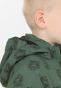 Groene jas met geometrische tijgerpatronen, voorzien van een capuchon, zwarte accenten en een ritsluiting. Glad stoffen textuur.