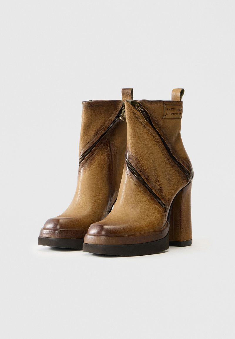 Bottines en cuir marron avec un haut talon carré, présentant des zips diagonaux à l'avant et une finition lisse. Accents texturés près des zips.