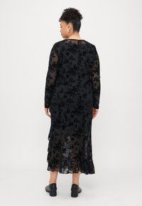 Robe en dentelle florale noire avec des manches longues, tissu transparent, ourlet en cascades et accents volants. Portée avec des chaussures à talons noires.