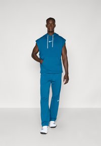 Blå ærmeløs hættebluse med frontlomme, hvide snore og Nike-logo, parret med matchende blå joggingbukser og hvide træningssko.