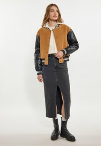 Giacca varsity in suede marrone e pelle nera con polsini a righe, abbinata a una gonna di denim nera con uno spacco laterale e stivali militari neri.