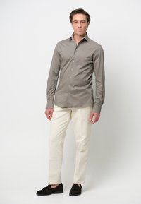 Camisa de botón de manga larga en gris con cuello puntiagudo, combinada con pantalones color crema y zapatos de ante oscuros sobre un fondo sencillo.