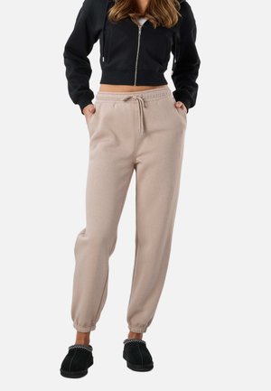 Kobieta w beżowych dresach ze ściągaczem, czarnej krótkiej bluzie z kapturem i suwakiem oraz czarnych wsuwanych butach, stoi z rękami w kieszeniach.