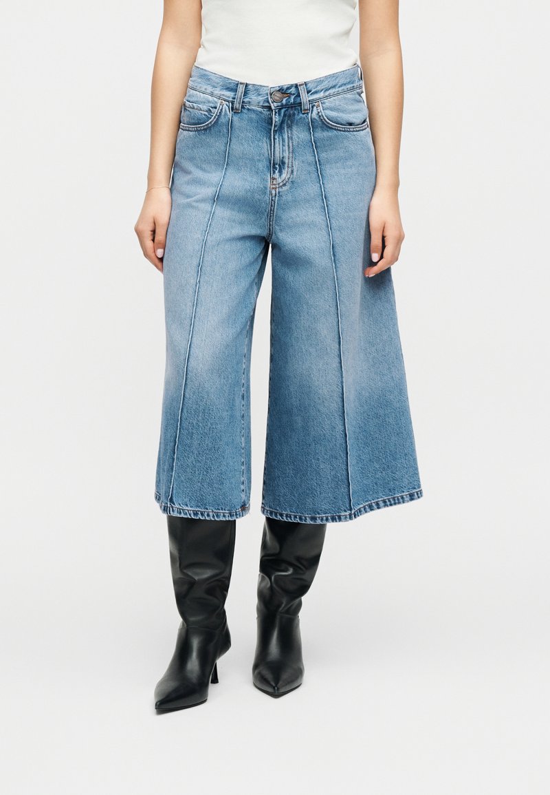 Mujer con jeans azules de pierna ancha y cortos, con costuras verticales visibles, botas negras de tacón hasta la rodilla y un top blanco sin mangas.