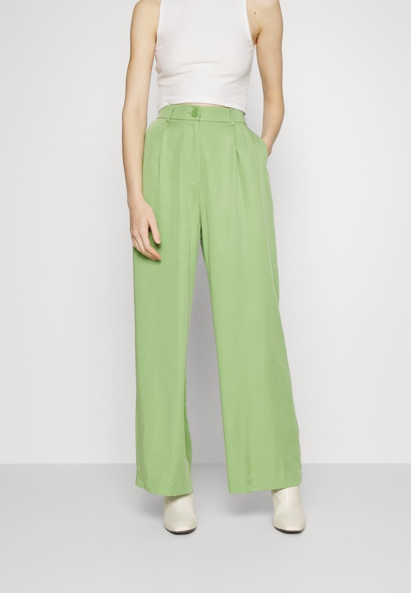 Pantalon large vert clair à taille haute, avec plis et fermeture à boutons. Associé à un crop top côtelé blanc et des chaussures neutres.