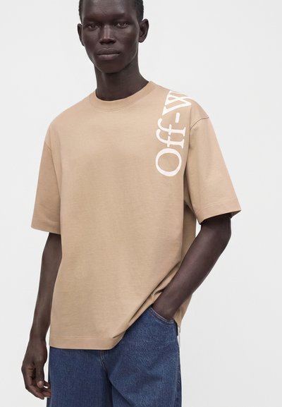 Homme portant un t-shirt beige Off-White avec un texte de logo blanc sur l'épaule, associé à un jean bleu large, debout devant un fond clair uni.