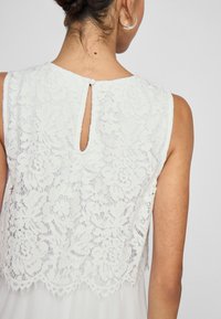 Hvit blondebluse med blomster mønster, med en nøkkelhullåpning bak og liten knappelukking. Glatt, lagdelt stoff.