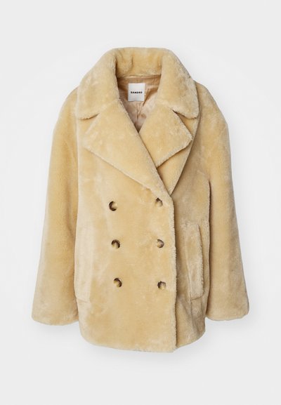 Beige Doppelreiher-Mantel aus Kunstfell mit Reverskragen, großen Knöpfen und seitlichen Taschen; weiche Textur und lockere Passform.