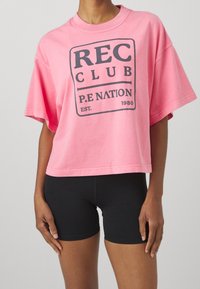 Růžové oversized bavlněné tričko s černým obdélníkovým logem s nápisem "REC CLUB P.E NATION EST. 1980," v kombinaci s černými sportovními šortkami.