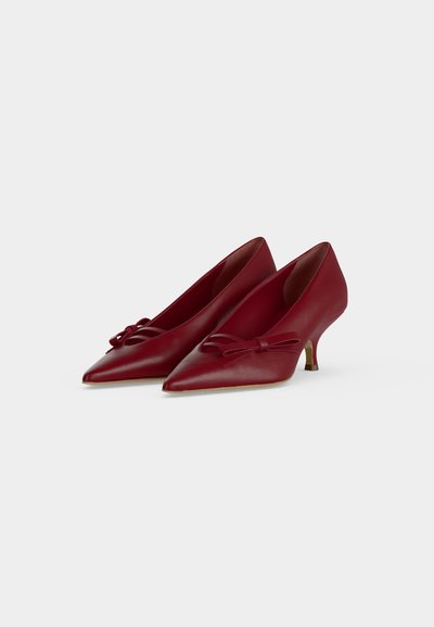 Paire d'escarpins rouges en cuir à bout pointu avec de petits nœuds sur le devant, sur un fond clair uni.