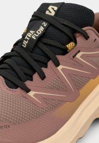 Sportsko med med teksturert rosa og beige overdel, svarte snøringer, gule detaljer og "Ultra Flow 2"-merkevaren på tungen.