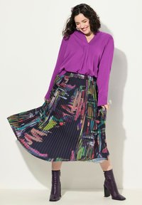 Blouse violette à manches longues associée à une jupe plissée multicolore arborant des motifs abstraits. Le mannequin porte des bottes à talons violettes.