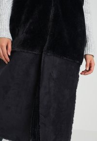 Černá vesta s kombinací umělé kožešiny a hladké látky. Má dlouhý, otevřený střih a hladké okraje bez švů. Viditelné jsou prošití detailů.