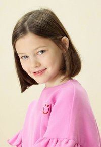 Sweatshirt rose avec des manches à volants et un visage souriant brodé en rouge. Le tissu semble doux et la coupe est décontractée.