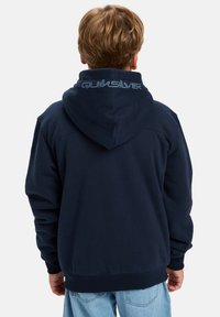 Tengerészkék kapucnis pulóver puha anyagból készült, nagy kapucnival és Quiksilver logóval a hátsó részén. Bordás mandzsetták és alja.