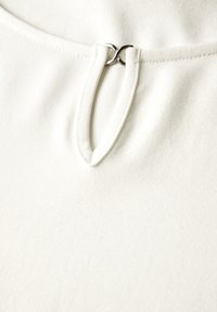 Top blanco con un escote en bucle y un acento metálico plateado, hecho de una tela suave con una textura lisa y un detalle de costura mínima.