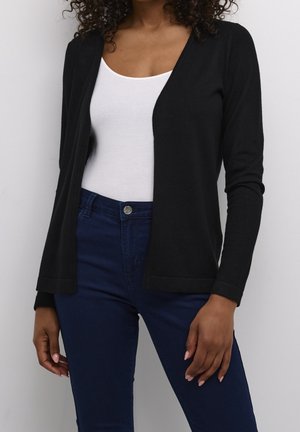 Femme portant un cardigan noir ouvert, un débardeur blanc et un jean bleu foncé, debout devant un fond clair uni.