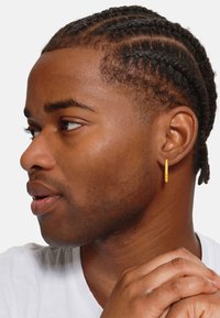 Goldene Creole mit einer glatten, zylindrischen Form; getragen von einer Person mit kurzen, geflochtenen Haaren; zeigt einen subtilen Glanz gegen den Hautton.