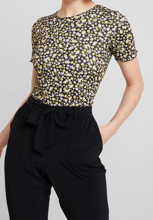 Vrouw draagt een zwart aansluitend kortärmelig shirt met kleine gele en witte bloemen, gecombineerd met zwarte hoge taillebroeken die in de taille zijn gestrikt.